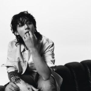 Black and white photo of Jungkook Calvin Klein from the brand’s campaign, seated on a dark sofa against a plain white background. He is wearing a T-shirt with the “Calvin Klein” logo partially visible, a light denim shirt open over it, and jeans. One hand is near his mouth in a thoughtful gesture, while the other rests on his leg. The tattoos on his right arm are prominently displayed, as well as his loose, wavy, slightly damp hair. Image published on the Bangtan Now blog.