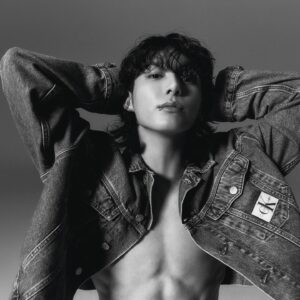 Black and white photo of Jungkook Calvin Klein from the brand’s campaign, set against a soft gray background. He is wearing an open Calvin Klein denim jacket, revealing his defined chest. His arms are raised with his hands behind his head, emphasizing a confident posture. His hair is loose and slightly wavy, with bangs covering part of his forehead. He looks directly at the camera with a calm expression and a visible lip piercing. Image published on the Bangtan Now blog.