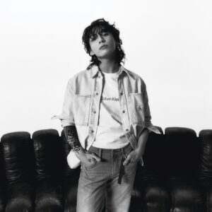 Black and white photo of Jungkook Calvin Klein from the brand’s campaign, featured on the Bangtan Now blog. He is standing in front of a dark leather sofa, wearing a white Calvin Klein T-shirt with the logo centered on the chest, a light denim shirt unbuttoned over it, and light-wash jeans with a visible belt detail. His left hand is in his pocket, and his right forearm displays a large tattoo. His wavy hair is loose, with bangs falling slightly over his eyes, and he looks at the camera with a calm, confident expression. The minimalist white background emphasizes the clean, modern aesthetic of the look.