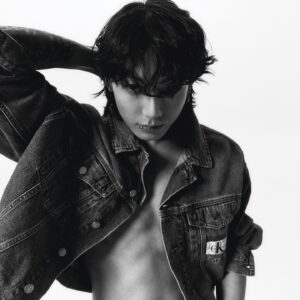 Black and white photo of Jungkook Calvin Klein from the brand’s campaign, set against a plain white background. He is wearing an open Calvin Klein denim jacket with no shirt underneath, revealing his bare torso. One hand is placed behind his head, highlighting the tattoo on his arm and creating a striking pose. His hair is loose and messy, with bangs partially covering his eyes. His gaze is intense and directed downward, with a visible lip piercing. Image published on the Bangtan Now blog.