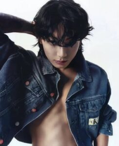 Color photo of Jungkook Calvin Klein from the brand’s campaign, set against a soft white background. He is wearing an open Calvin Klein denim jacket, exposing his chest. With one hand placed behind his head, he strikes a bold pose that highlights the tattoo on his left arm. His black hair is loose, with bangs falling over his eyes, and his lips have a slight shine under the light. His gaze is intense and directed downward. Image published on the Bangtan Now blog.