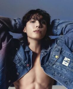 Jungkook Calvin Klein em foto colorida da campanha da marca, com fundo azul suave. Ele aparece de perto, com expressão confiante, cabelo solto com franja cobrindo parte da testa e um piercing no lábio inferior. Jungkook veste jaqueta jeans Calvin Klein aberta, deixando o peitoral definido à mostra. Seus braços estão levantados, com as mãos apoiadas atrás da cabeça. A jaqueta exibe o logo da marca no bolso e botões personalizados. Imagem publicada no blog Bangtan Now.