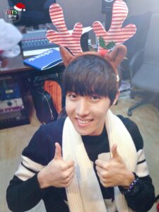 Na imagem, j-hope aparece sorrindo amplamente enquanto faz sinal de positivo com as duas mãos, usando uma tiara de rena listrada em vermelho e branco e um cachecol branco que completa o look festivo. Ele está em um estúdio, com equipamentos de áudio ao fundo, papéis espalhados e uma pasta azul da Adidas sobre a mesa, o que reforça o clima casual e espontâneo dos bastidores. A iluminação natural suaviza a cena e destaca o brilho nos olhos dele, transmitindo a alegria genuína que marcou o especial natalino "[FM 06.13] The very happy Christmas with BTS!". Essa imagem é mais uma daquelas que mostram como o BTS Natal sempre foi construído com simplicidade e afeto, encantando o ARMY com pequenos gestos e muito carisma. Resgatada pelo Bangtan Now, ela representa bem como os membros, mesmo nos momentos mais descontraídos, conseguem aquecer o coração de quem acompanha sua trajetória.