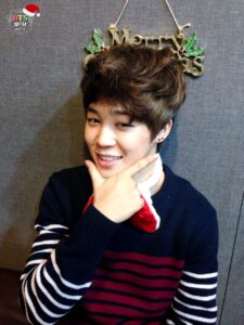 Na imagem, Jimin aparece sorrindo com um olhar brincalhão, fazendo uma pose clássica com a mão sob o queixo, como se estivesse se apresentando para um ensaio divertido. Ele veste um suéter listrado em azul-marinho, vermelho e branco, com um pequeno detalhe de acessório natalino no pescoço. Atrás dele, pendurado na parede, está um enfeite dourado escrito "Merry Christmas", com folhas de azevinho decorativas, completando o clima festivo. A parede cinza do estúdio funciona como fundo neutro, dando destaque ao sorriso e ao carisma natural do Jimin, em mais um registro do especial "[FM 06.13] The very happy Christmas with BTS!". Essa imagem transmite leveza e aquele charme espontâneo dos primeiros anos do grupo, que conquistava fãs pela naturalidade e pela conexão sincera. É mais um retrato caloroso do BTS Natal, recuperado com carinho pelo Bangtan Now para celebrar essa época tão especial com quem carrega o Bangtan no coração.