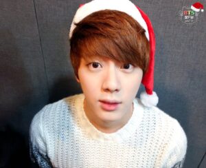 Na imagem, Jin aparece usando um gorro de Natal vermelho com detalhes brancos e um suéter de tricô claro, olhando diretamente para a câmera com uma expressão curiosa e cativante. A parede cinza do estúdio de rádio do especial "[FM 06.13] The very happy Christmas with BTS!" serve como plano de fundo, com o logo no canto superior direito reforçando o clima natalino. A iluminação suave destaca seus traços delicados e o brilho nos olhos que conquistam qualquer ARMY à primeira vista. Essa foto é mais um dos registros preciosos do BTS Natal, resgatado com carinho pelo Bangtan Now para aquecer a memória afetiva de quem acompanha o grupo desde o comecinho.