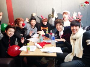 Na imagem, os sete membros do BTS aparecem reunidos ao redor de uma mesa no especial natalino "[FM 06.13] The very happy Christmas with BTS!", todos sorrindo e fazendo poses animadas em um momento que transborda energia e alegria juvenil. Cada integrante veste acessórios natalinos como gorros de Papai Noel, tiaras de rena, chapéus com mensagens festivas e até nariz de rena, criando um clima descontraído e afetuoso. Sobre a mesa há papéis com anotações, bebidas e uma meia de Natal, compondo um cenário simples, mas cheio de personalidade. A parede ao fundo tem um pequeno letreiro com "Merry Christmas", e o logo do programa aparece no canto superior direito. Essa foto captura o início da conexão calorosa entre o grupo e os fãs, e simboliza o espírito divertido e acolhedor que marca o BTS Natal desde os primeiros anos. Resgatada pelo Bangtan Now, essa imagem é um registro especial que mostra o BTS como o ARMY aprendeu a amar: espontâneo, unido e cheio de carisma mesmo nas produções mais simples.