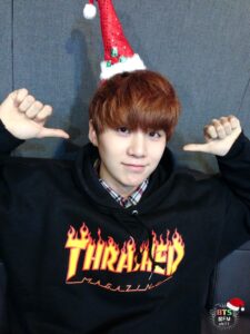 Na imagem, Yoongi aparece com um mini chapéu de Natal vermelho com lantejoulas, decorado com detalhes brancos e verdes, equilibrado no topo da cabeça de forma divertida. Ele veste um moletom preto com a logo da Thrasher Magazine em laranja e vermelho flamejante, e por baixo, uma camisa xadrez que adiciona um toque casual ao look. Sorrindo levemente e apontando com os polegares para si mesmo, ele transmite aquele charme discreto e inconfundível que só o SUGA tem. A parede cinza e o logo do especial "[FM 06.13]" no canto superior direito completam a ambientação do programa de rádio natalino do grupo. É mais uma lembrança especial do BTS Natal resgatada pelo Bangtan Now, perfeita para aquecer o coração dos fãs nessa época do ano.