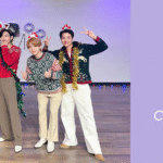 Imagem promocional publicada pela BigHit no X (antigo Twitter) como parte da mensagem de Happy Holidays, usada como capa do artigo especial sobre BTS Natal no blog do Bangtan Now. No lado esquerdo da imagem, os sete membros do BTS aparecem juntos em um ambiente interno decorado para o fim de ano. Eles vestem suéteres com estampas natalinas, cada um em um estilo diferente, e acessórios festivos como tiaras de rena, orelhinhas de coelho e pequenos chapéus de Papai Noel. As poses são descontraídas, com sorrisos, gestos de paz e muita leveza, transmitindo uma energia acolhedora e alegre. Ao fundo, balões metálicos coloridos formam a frase "HAPPY HOLIDAY", acompanhados por luzes pisca-pisca discretas que reforçam o clima natalino. O cenário é simples, mas carismático, com piso de madeira clara e iluminação suave. À direita da imagem, sobre um fundo lilás, está o título do artigo: "BTS NATAL – Músicas, vídeos e fotos para celebrar as festas com o Bangtan", junto ao logotipo do Bangtan Now, destacando o conteúdo especial dedicado ao ARMY. Esta imagem resume perfeitamente o espírito do BTS Natal. É visualmente acessível, afetiva e feita para despertar aquele sentimento bom de fim de ano com o Bangtan no coração.