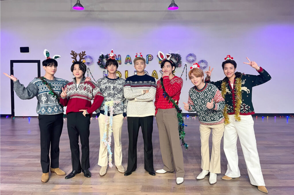 Em uma nova pose diante do mesmo cenário festivo, os integrantes do BTS aparecem sorridentes com trajes natalinos coloridos, adornados com acessórios divertidos como orelhas de coelho e chapéus de Papai Noel, reforçando a energia leve e animada do BTS Natal. Publicada originalmente no X, a foto integra o especial do blog Bangtan Now.