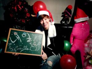 Na imagem, Jimin aparece sentado sorridente em um ambiente decorado para o Natal, vestindo um gorro de Papai Noel e um cachecol branco, com um visual casual e cheio de charme. Ele segura uma lousa verde com desenhos natalinos feitos à mão em giz branco, incluindo um boneco de neve, corações e pequenas mensagens escritas em coreano. Ao seu redor, há balões vermelhos e verdes, uma guirlanda decorada e, claro, o famoso boneco de pelúcia rosa com gorro de Natal que aparece em outras fotos do grupo. Ao fundo, também é possível ver uma árvore branca decorada e um ambiente com iluminação suave que dá um tom acolhedor à cena. Publicada no perfil oficial do BTS no X, essa foto é um exemplo perfeito da forma carinhosa e próxima como os meninos celebravam essa época com os fãs. É um registro simples, mas cheio de significado, que carrega o sentimento afetivo do BTS Natal, resgatado com cuidado pelo Bangtan Now para reviver memórias inesquecíveis entre ARMYs.
