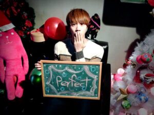 Na imagem, Jin está sentado em uma poltrona escura, cercado por decorações natalinas, segurando uma lousa com a palavra "Perfect" escrita à mão em giz azul, com desenhos de estrelinhas ao redor. Ele leva uma das mãos à boca em uma expressão surpresa ou brincalhona, e veste um suéter de inverno em tons claros. À sua volta, há uma árvore de Natal branca decorada com bolas coloridas e laços dourados, balões vermelhos e verdes, presentes embrulhados e o mesmo boneco de pelúcia rosa que aparece em outras fotos natalinas do grupo. O ambiente tem uma iluminação suave e aconchegante, com sombras que reforçam a sensação de cena montada com carinho. Essa imagem foi publicada no perfil oficial do BTS no X durante uma das celebrações de fim de ano e se tornou mais um dos registros adorados pelo ARMY. É uma cena que resume bem o clima leve, divertido e afetuoso do BTS Natal, e que o Bangtan Now resgata como parte dessas memórias que merecem ser revisitadas.