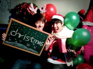 Na imagem, Jung Kook e j-hope aparecem sentados lado a lado em um ambiente decorado para o Natal, transmitindo alegria e espontaneidade. Jung Kook segura uma lousa com a palavra "Christmas" escrita em giz branco, sorrindo enquanto usa uma tiara de rena com detalhes vermelhos. j-hope está ao seu lado com um gorro de Papai Noel e um cachecol branco volumoso, segurando balões verdes e fazendo uma expressão animada com as mãos formando um coração. Ao fundo, há balões vermelhos e verdes, guirlanda, e o famoso boneco de pelúcia rosa já conhecido de outras fotos natalinas do grupo, reforçando o clima de brincadeira entre os membros. A iluminação suave dá um ar acolhedor e afetuoso, bem característico dos primeiros conteúdos festivos do grupo. Essa foto foi publicada no perfil oficial do BTS no X e representa perfeitamente o espírito leve, divertido e caloroso do BTS Natal, sendo mais um dos registros especiais resgatados com carinho pelo Bangtan Now.