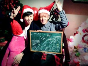Na imagem, RM e Taehyung aparecem sentados lado a lado, usando gorros de Papai Noel e fazendo caretas divertidas enquanto levantam os braços em uma pose brincalhona. Entre eles, seguram uma lousa com desenhos feitos à mão em giz branco, com a data “2013.12.18” escrita em destaque, marcando um dos primeiros registros festivos do grupo. À esquerda, o já familiar boneco de pelúcia rosa com gorro natalino aparece novamente, reforçando a identidade visual desse conjunto de imagens. Ao fundo, balões vermelhos e verdes, uma guirlanda e uma árvore de Natal branca decorada com bolas coloridas completam o cenário acolhedor e lúdico. A atmosfera da foto é leve e cheia de charme caseiro, capturando o entusiasmo jovem e sincero dos primeiros anos do BTS. Publicada no perfil oficial do grupo no X, essa imagem é um retrato carinhoso do espírito do BTS Natal, e mais uma memória preciosa resgatada com cuidado pelo Bangtan Now para aquecer o coração do ARMY.