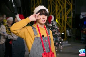 Em mais um momento adorável do Run BTS! Ep. 32 – Please Take Care of Santa (Pigdolf), Jungkook aparece com um sorriso tímido e fazendo o clássico “V” de paz. Ele veste um moletom amarelo vibrante por baixo de uma jardineira listrada, acompanhado de um gorro de Papai Noel e a bolsinha vermelha característica do episódio. Ao fundo, o cenário natalino ganha vida com uma árvore decorada e outros membros interagindo, criando uma atmosfera festiva e divertida. Um registro que combina perfeitamente com o espírito de BTS Natal e o encanto do Bangtan Now.