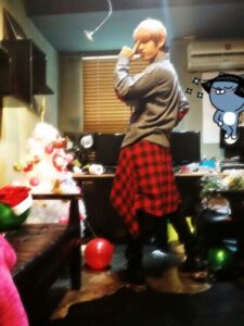 Na imagem, Taehyung (V) aparece em uma pose divertida e cheia de personalidade, com um olhar marcante e os dedos fazendo o clássico “V” de vitória ao lado do rosto. Ele veste um suéter cinza com uma camisa xadrez vermelha amarrada na cintura, em um look casual com aquele toque único que só ele tem. O cenário ao fundo é uma sala simples e bagunçada, com mesas cheias de objetos, cabos, papéis e computadores, contrastando com os elementos natalinos espalhados pelo ambiente. À esquerda, uma pequena árvore de Natal branca decorada com bolas coloridas chama atenção, além de balões vermelhos e verdes espalhados pelo chão. A iluminação quente e a decoração criam um clima aconchegante e despretensioso. A imagem foi publicada no perfil oficial do BTS no X durante a época de festas, e representa bem o espírito espontâneo e divertido do BTS Natal. Esse tipo de registro, resgatado com carinho pelo Bangtan Now, reforça como até os momentos mais simples têm o poder de virar memória afetiva entre os fãs.