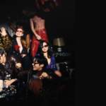 Cover for the article "NORMAL BTS Lyrics: Meaning Behind Every Verse" from the Bangtan Now blog, with the Articles category tag and logo. Photo in dark tones with a vintage party atmosphere: the seven BTS members pose in colorful, eye-catching outfits, including leopard print, sequins, and one person in a long red dress standing on a sofa, with relaxed gestures like waving and leaning on each other, gathered around a table with a cake
