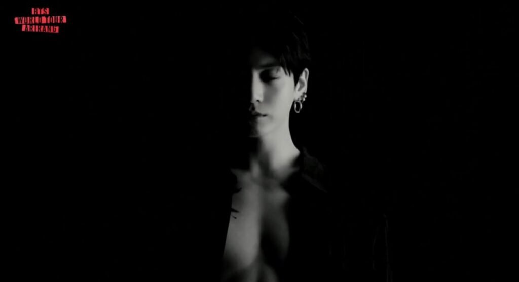 Retrato em preto e branco de Jung Kook envolto em escuridão, rosto e ombro parcialmente iluminados por luz lateral suave, olhos fechados, brincos visíveis na orelha, com o logo "BTS WORLD TOUR ARIRANG" no canto superior esquerdo, no VCR dos sete elementos da turnê.