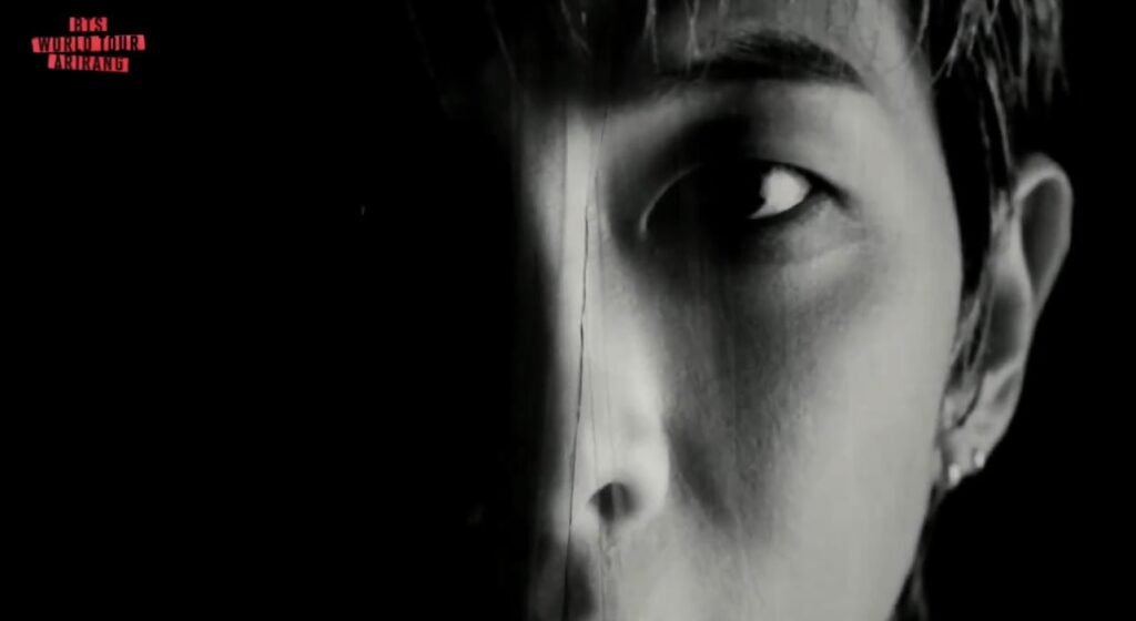 Close extremo do rosto de Namjoon em preto e branco, metade do rosto na sombra, olho fixando a câmera, cabelo molhado, com o logo "BTS WORLD TOUR ARIRANG" no canto superior esquerdo, no VCR dos sete elementos da turnê.