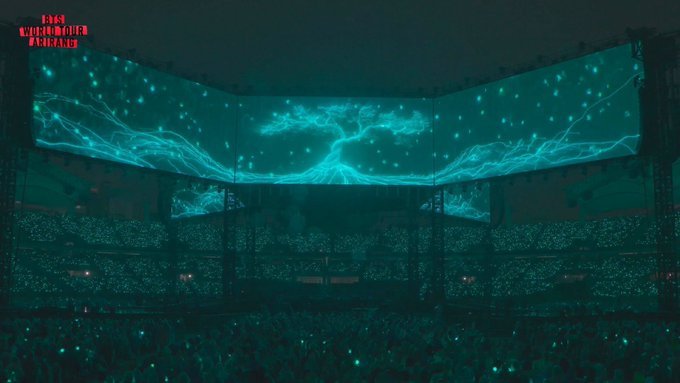 Telões do palco da turnê ARIRANG do BTS exibindo, em tons de verde-azulado brilhante contra o céu escuro, uma árvore luminosa se formando a partir de raízes ramificadas como raios, a Yeonriji do segundo VCR do show. Abaixo, o estádio lotado com luzes de celular acesas, e o logo "BTS WORLD TOUR ARIRANG" no canto superior esquerdo.