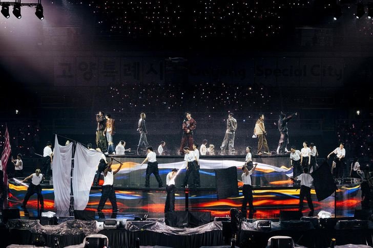 Referência ao Ganggangsullae no palco da turnê ARIRANG do BTS: dançarinos vestidos de branco seguram tecidos coloridos formando ondas na base do palco, enquanto os sete integrantes do BTS caminham em fila sobre a plataforma elevada, sob chuva de partículas de luz. Ao fundo, uma faixa com os dizeres "고양특례시 Goyang Special City".