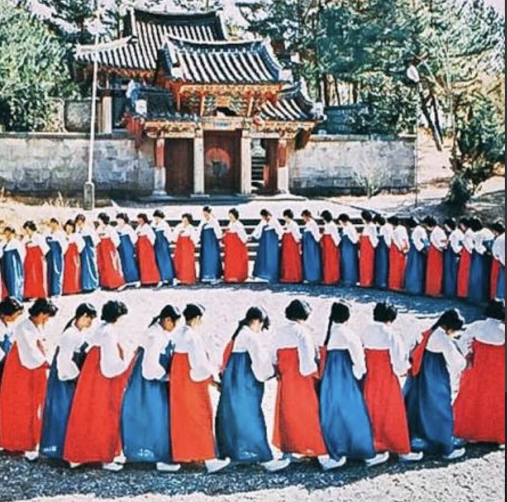 Mulheres em hanbok vermelho e azul, cabelo preso em coques tradicionais, dançam o Ganggangsullae, dança folclórica coreana do festival da colheita, formando círculos concêntricos de mãos dadas em um pátio de terra batida. Ao fundo, um portão tradicional coreano de telhas escuras e vermelho, cercado por árvores.