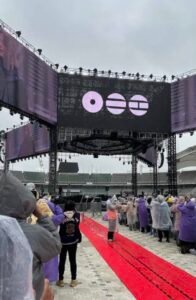 O palco central da turnê ARIRANG do BTS, com estrutura elevada de treliças metálicas em formato que remete ao jeongja e telões exibindo três símbolos circulares roxos, o logo da turnê. Em primeiro plano, uma mão segura um leque preto redondo com a inscrição "ARIRANG WORLD TOUR". Ao fundo, arquibancadas vazias de um estádio sob céu nublado.