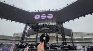 O palco central da turnê ARIRANG do BTS, com estrutura elevada de treliças metálicas em formato que remete ao jeongja e telões exibindo três símbolos circulares roxos, o logo da turnê. Em primeiro plano, uma mão segura um leque preto redondo com a inscrição "ARIRANG WORLD TOUR". Ao fundo, arquibancadas vazias de um estádio sob céu nublado.