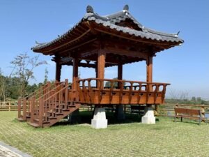Um jeongja tradicional coreano, pavilhão de madeira elevado sobre pilares de pedra, com telhado de telhas cinzas em beiral curvo e escada de acesso à plataforma - a estrutura que inspirou o palco da turnê ARIRANG do BTS.