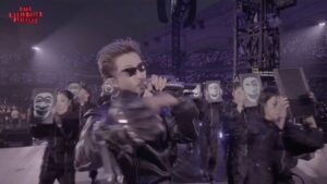 Namjoon canta ao microfone no palco da turnê ARIRANG do BTS, cabelo loiro e óculos escuros, vestindo jaqueta de couro preta. Ao fundo, dançarinos seguram telas exibindo a mesma máscara tradicional coreana (tal), e o estádio lotado aparece coberto de luzes de fã, com o logo "BTS WORLD TOUR ARIRANG" no canto superior esquerdo.