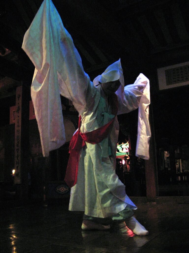 Um dançarino da dança seungmu, tradição budista coreana, gira com os braços erguidos usando um capuz branco e mangas longas que se estendem no ar, uma faixa vermelha amarrada na cintura. Ao fundo, um templo com lanternas acesas na escuridão. Essa dança inspirou a coreografia de "Merry Go Round" na turnê ARIRANG do BTS.