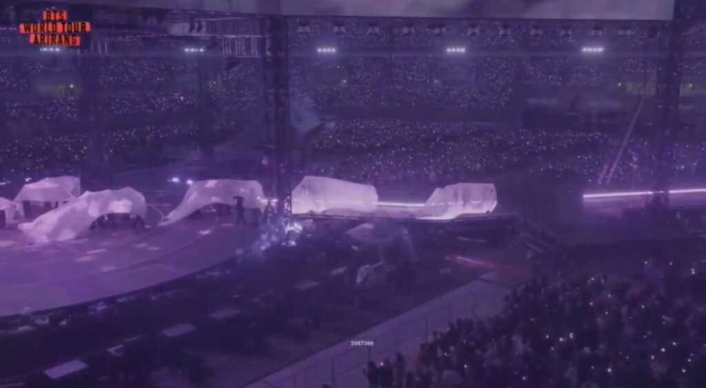 Vista panorâmica do palco da turnê ARIRANG do BTS coberto por grandes tecidos brancos drapeados, sob luz roxa, remetendo às mangas da dança seungmu do momento de "Merry Go Round". Ao redor, o estádio lotado com o público erguendo luzes de celular, e o logo "BTS WORLD TOUR ARIRANG" no canto superior esquerdo.