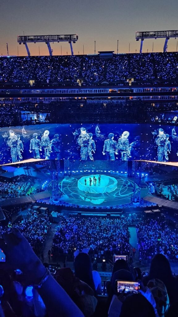 Vista de dentro do público durante "SWIM" na turnê ARIRANG do BTS, todo o estádio banhado em luz azul. No centro, os sete integrantes do BTS em silhueta na base circular do palco, cercados por tecidos brancos ondulantes que imitam água. Os telões ao redor projetam a mesma cena ampliada, e a plateia lotada segura celulares e luzes acesas em primeiro plano.