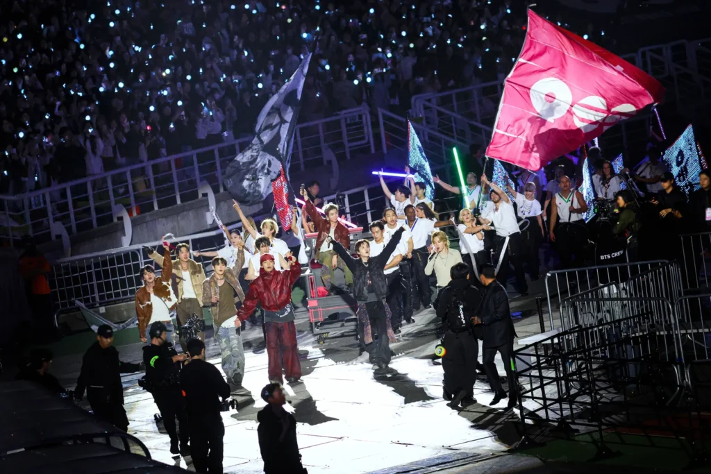 A procissão de "IDOL" na turnê ARIRANG do BTS: integrantes do BTS e dançarinos caminham pela pista ao redor do estádio, de braços erguidos, cercados por bandeiras vermelhas e pretas com o símbolo da turnê e fitas de LED em azul e verde. Ao fundo, o público lotado balança luzes de celular no escuro.