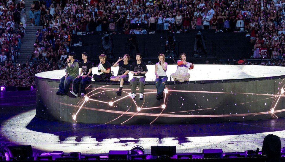 Os sete integrantes do BTS sentados na base circular do palco da turnê ARIRANG do BTS, durante "Please", vestidos em tons de cinza e branco. Abaixo deles, a base exibe a projeção da constelação da Ursa Maior em traços dourados. Ao fundo, o público lotado acena luzes de celular sob luz roxa.