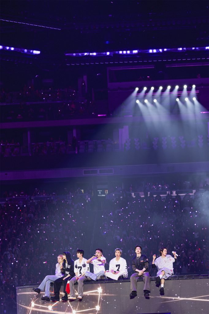 Os sete integrantes do BTS sentados na base circular do palco da turnê ARIRANG do BTS, durante "Please", em roupas casuais nas cores verde, preto e bege. A base exibe a projeção da constelação da Ursa Maior em traços claros. Ao fundo, o público lotado sob luz branca intensa.