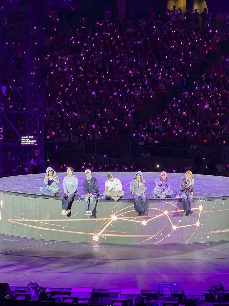 Os sete integrantes do BTS sentados lado a lado na base elevada e circular do palco da turnê ARIRANG do BTS, durante "Please", vestindo peças brancas e alguns com o número 7. Ao fundo, o estádio lotado sob luz roxa, com holofotes cruzando o alto do palco.