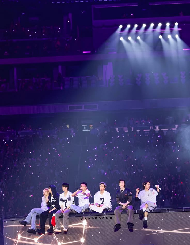 Vista de longe dos sete integrantes do BTS sentados na base do palco da turnê ARIRANG do BTS, durante "Please", em roupas brancas e pretas com o número 7. O estádio aparece em múltiplos andares lotados, com holofotes brancos iluminando o alto do palco.