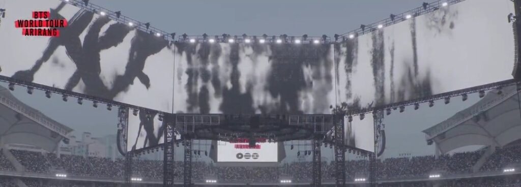 Telão do palco da turnê ARIRANG do BTS exibindo sete silhuetas escuras em efeito de wash painting (sumukhwa), como figuras humanas se dissolvendo em tinta preta diluída sobre fundo branco, sob as luzes de palco no topo da tela.