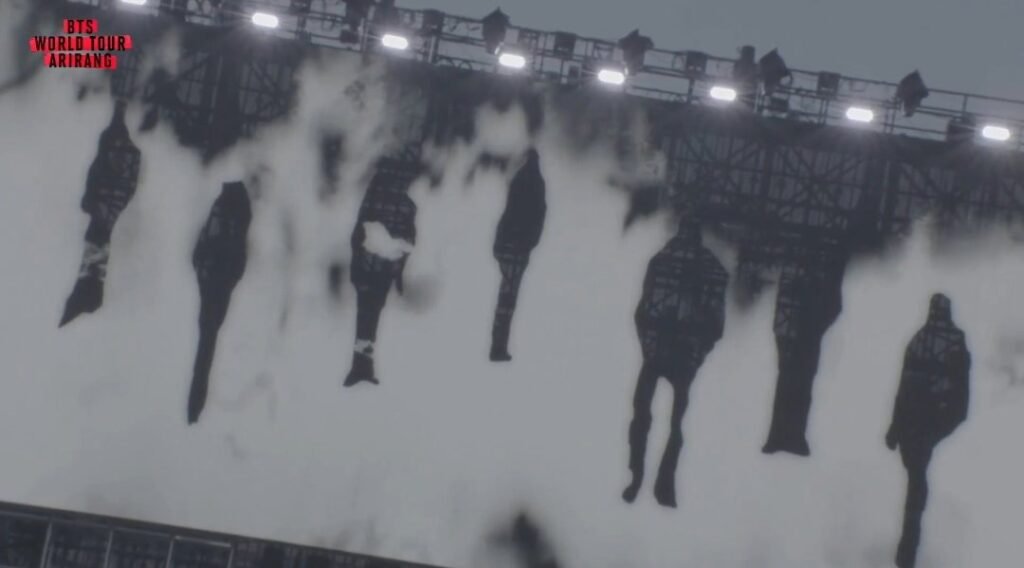 Vista aberta do palco da turnê ARIRANG do BTS, com os telões ao redor de toda a estrutura exibindo o mesmo efeito de wash painting em preto e branco, silhuetas dissolvendo-se como tinta sobre papel. Ao fundo, o telão central menor mostra o logo da turnê e o símbolo circular do jeongja, com o estádio lotado ao redor sob céu nublado.