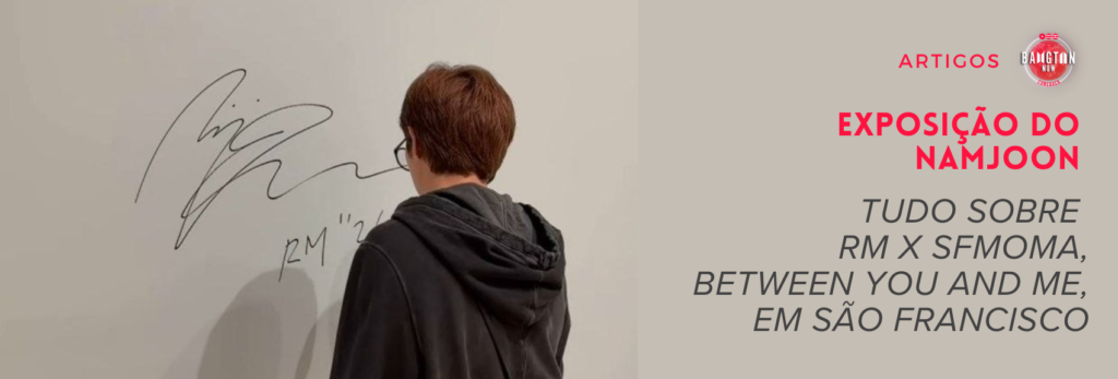 Exposição do Namjoon no SFMOMA: o Namjoon aparece de costas, com o cabelo curto castanho-avermelhado, óculos e um moletom com capuz cinza-escuro, diante de uma parede clara do museu onde acabou de deixar uma grande assinatura em traço preto, com "RM" e uma data parcialmente encoberta pela cabeça dele. A foto foi compartilhada pelo próprio Namjoon nos stories do Instagram. À direita, sobre fundo bege, a capa do Bangtan Now traz o logo do blog, a palavra "Artigos" e os textos "Exposição do Namjoon", em vermelho, e "Tudo sobre RM x SFMOMA, Between You and Me, em São Francisco", em cinza.