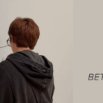 Exposição do Namjoon no SFMOMA: o Namjoon aparece de costas, com o cabelo curto castanho-avermelhado, óculos e um moletom com capuz cinza-escuro, diante de uma parede clara do museu onde acabou de deixar uma grande assinatura em traço preto, com "RM" e uma data parcialmente encoberta pela cabeça dele. A foto foi compartilhada pelo próprio Namjoon nos stories do Instagram. À direita, sobre fundo bege, a capa do Bangtan Now traz o logo do blog, a palavra "Artigos" e os textos "Exposição do Namjoon", em vermelho, e "Tudo sobre RM x SFMOMA, Between You and Me, em São Francisco", em cinza.