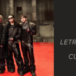 Banner de portada del artículo sobre la letra y traducción de Hooligan BTS. A la izquierda, los siete integrantes de BTS en el photo sketch de Hooligan, publicado en Weverse, posan uno al lado del otro sobre un piso rojo brillante, en un galpón industrial de concreto gris con columnas altas y reflectores al fondo. Todos visten looks negros de cuero, con chaquetas, pantalones anchos, botas pesadas, cadenas plateadas y pañuelos estampados al cuello; uno hace el símbolo de la paz con la mano y otro usa lentes oscuros y chaqueta con cuello de piel. A la derecha, sobre fondo gris oscuro, aparecen el logo de Bangtan Now, la palabra "Artículos" y el texto: "Traducción de Hooligan: letra, pronunciación, qué significa y curiosidades que ni te imaginabas."